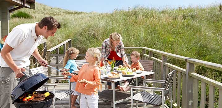 Familie på en terrasse ved et sommerhus. Far serverer for datter fra grillen, mens resten af familien sidder ved bordet i baggrunden